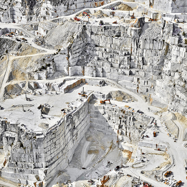 An aerial view of Quarry 05, Carrara, IT, 2018, by David Burdeny shows terraced formations of light-colored stone blocks. Construction vehicles navigate the winding paths amidst ongoing environmental debates about extraction processes.