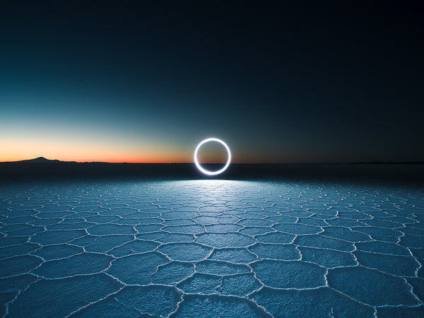 The XT1876 by Reuben Wu creates a luminous circle, likely from light-carrying drones, above Salar de Uyunis vast, cracked salt flats at dawn or dusk. The sky shifts from deep blue to orange near the horizon, stars sprinkled subtly across it. Geometric patterns add texture to the serene scene.